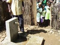 Un adulte ouvre l'eau de la nouvelle borne-fontaine, devant le regard des enfants, se tenant respectuant à l'entrée de l'enclos 